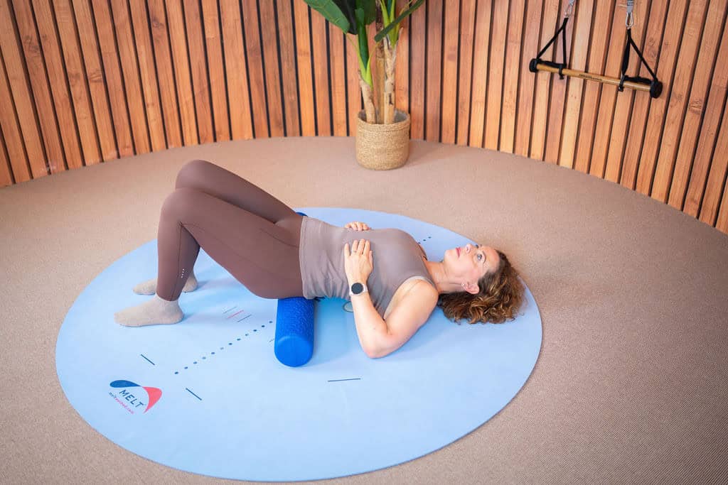 Vrouw ligt op een blauwe MELT-mat met foam roller onder haar knieën.
